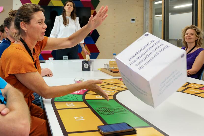 Een groep mensen zit aan een tafel. Een vrouw gooit een grote dobbelsteen.